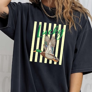 May include: Dark navy blue t-shirt featuring a graphic of a flying duck with green, brown, and blue feathers. The word "Lucky" is written in green script above the duck, set against a background of yellow and white vertical stripes.