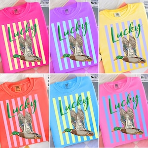 May include: Crew neck t-shirts in various colors, each featuring a graphic of a flying duck with the word "Lucky" in green script. The design includes vertical stripes in contrasting colors.