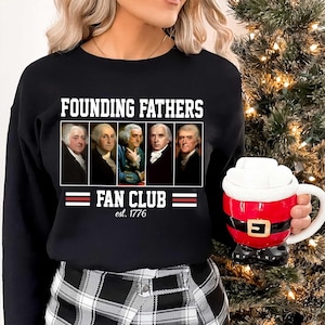 Może przedstawiać: Czarna bluza z napisem "FOUNDING FATHERS FAN CLUB est. 1776" nad rzędem portretów. Czerwono-biały kubek Świętego Mikołaja trzymany jest w prawej dłoni.