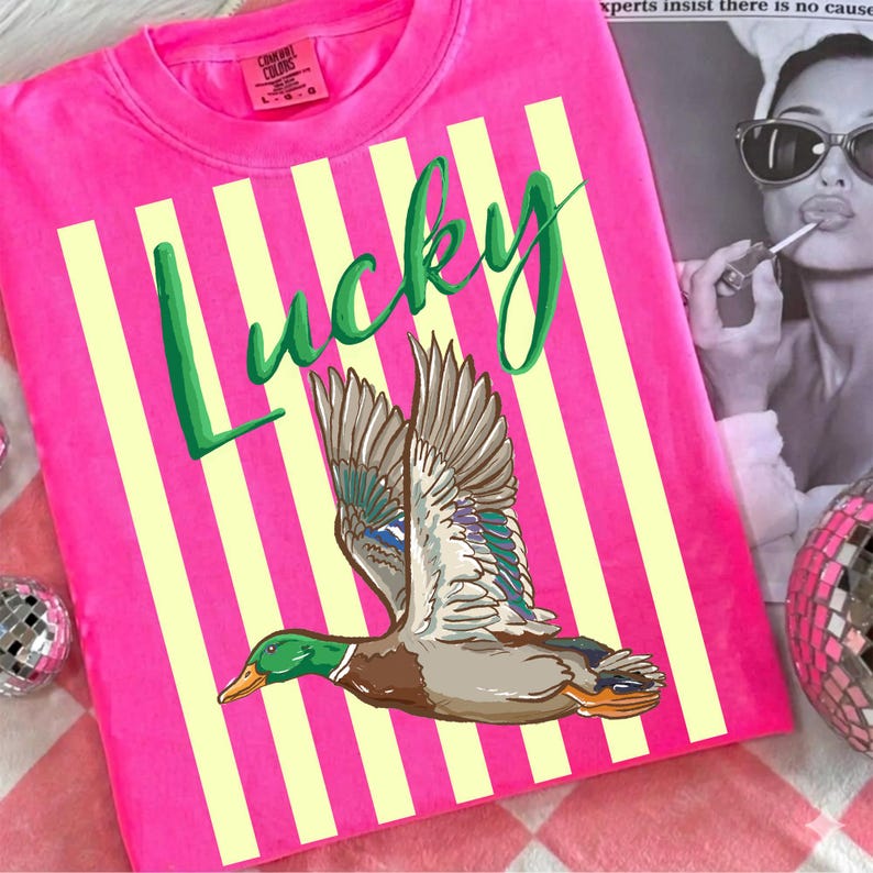 May include: A bright pink t-shirt with vertical yellow stripes and the word "Lucky" in green script. A detailed illustration of a flying duck with brown, green, and blue feathers is featured on the shirt.