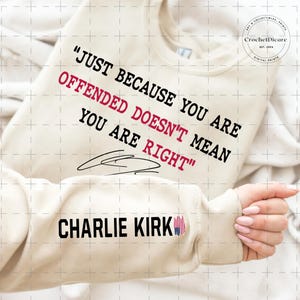 May include: Cream-colored sweatshirt with the text "JUST BECAUSE YOU ARE OFFENDED DOESN'T MEAN YOU ARE RIGHT" in black and red. The sleeve features "CHARLIE KIRK" in black with a small American flag graphic. The sweatshirt is a casual apparel item.
