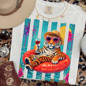 May include: White t-shirt featuring a tiger wearing sunglasses and a straw hat, relaxing in a red inflatable ring and holding a glass of red wine. The shirt design includes a tropical beach scene with palm trees and a yellow sun.