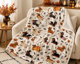 May include: A cream-colored throw blanket featuring a repeating pattern of dachshunds in various Halloween costumes, including pumpkins, ghosts, and witch hats, interspersed with small spiders, webs, bats, and candy icons on a soft fabric.