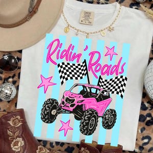Peut inclure: T-shirt blanc avec un dessin d'un v&eacute;hicule tout-terrain rose et l'inscription "Ridin' Roads" en rose. Le motif comprend des drapeaux &agrave; damiers et des &eacute;toiles roses. Le t-shirt a un col rond et des manches courtes.