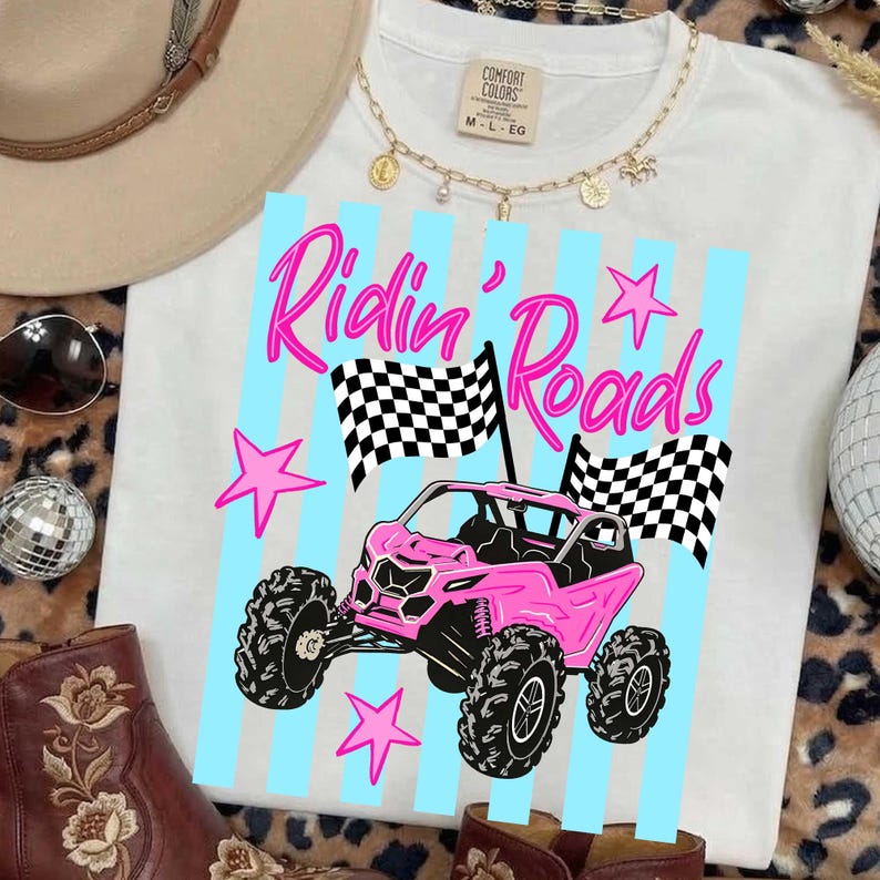 Peut inclure: T-shirt blanc avec un dessin d'un v&eacute;hicule tout-terrain rose et l'inscription "Ridin' Roads" en rose. Le motif comprend des drapeaux &agrave; damiers et des &eacute;toiles roses. Le t-shirt a un col rond et des manches courtes.