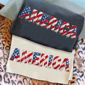 Peut inclure: Deux sweatshirts, un bleu marine et un crème, sont présentés. Chacun affiche le mot "AMERICA" dans un design patriotique, avec des lettres faites de morceaux du drapeau américain. Les sweatshirts sont pliés et empilés.