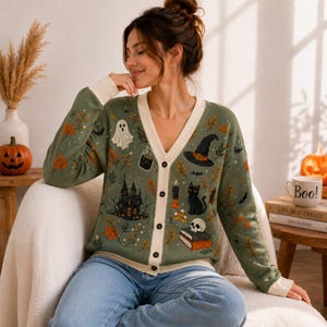 May include: A sage green V-neck button-down cardigan featuring a Halloween-themed print with a haunted house, a ghost, a black cat, a witch hat, a skull on books, a candle, and autumn leaves. The sweater has a cream-colored placket and cuffs.