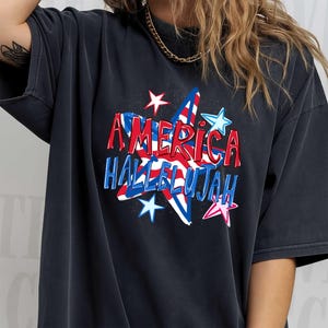 May include: Dark gray t-shirt featuring the words "AMERICA HALLELUJAH" in red and blue, with a star design. The design includes red and white stripes and blue stars. The shirt has short sleeves.