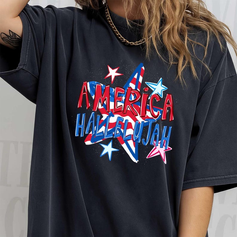 May include: Dark gray t-shirt featuring the words "AMERICA HALLELUJAH" in red and blue, with a star design. The design includes red and white stripes and blue stars. The shirt has short sleeves.