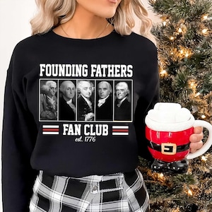 Może przedstawiać: Czarna bluza z napisem "FOUNDING FATHERS FAN CLUB est. 1776" i portretami postaci historycznych. W dłoni trzymany jest czerwono-biały kubek z motywem Świętego Mikołaja.