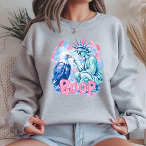 May include: A light grey sweatshirt with a graphic of an eagle and the Statue of Liberty. The eagle is blue and white, and the statue is turquoise. The word "BOOP" is in pink, with a blue and pink background.