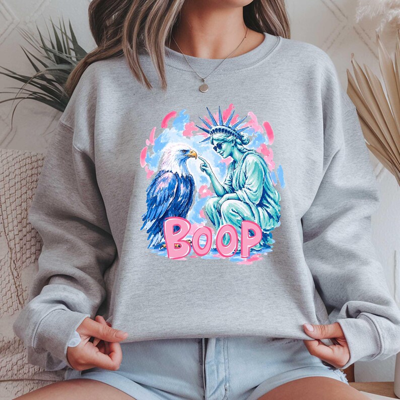 May include: A light grey sweatshirt with a graphic of an eagle and the Statue of Liberty. The eagle is blue and white, and the statue is turquoise. The word "BOOP" is in pink, with a blue and pink background.