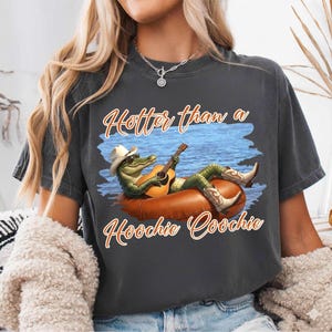 Peut inclure: T-shirt gris foncé avec un alligator de dessin animé portant un chapeau et des bottes de cow-boy, jouant de la guitare sur un bouée. Le texte dit "Hotter than a Hoochie Coochie."