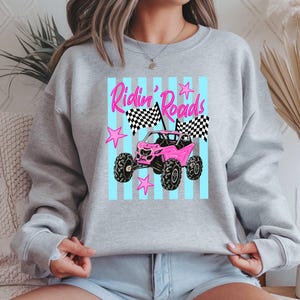 Peut inclure: Un sweat-shirt gris clair avec un motif de v&eacute;hicule tout-terrain rose et le texte "Ridin' Roads". Le motif comprend des drapeaux &agrave; damier et des &eacute;toiles roses sur un fond &agrave; rayures bleu clair.