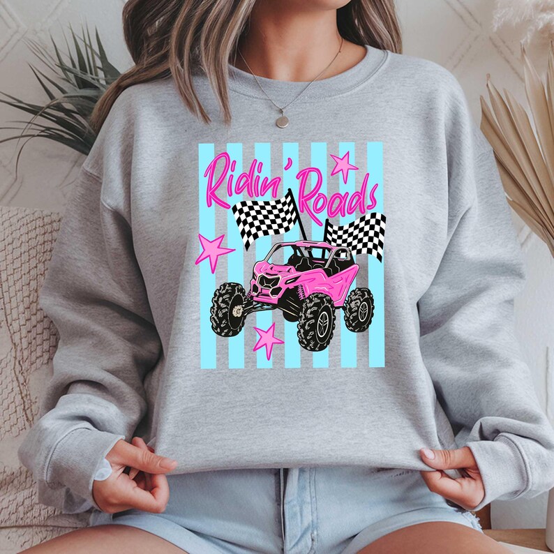 Peut inclure: Un sweat-shirt gris clair avec un motif de v&eacute;hicule tout-terrain rose et le texte "Ridin' Roads". Le motif comprend des drapeaux &agrave; damier et des &eacute;toiles roses sur un fond &agrave; rayures bleu clair.