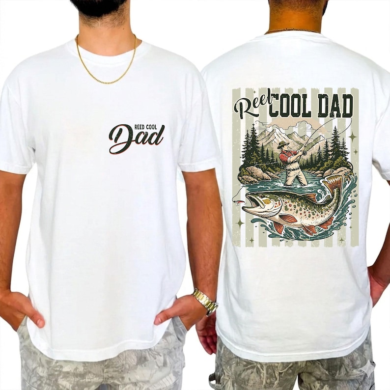 May include: White t-shirt with a "Reel Cool Dad" graphic on the back, depicting a fisherman, fish, and mountain landscape. The front displays "Reel Cool Dad" in script. A relaxed, comfortable tee.