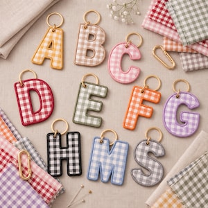 Könnte beinhalten: Eine Sammlung von Stoff-Schlüsselanhängern in Buchstabenform mit Vichy-Karo-Muster in verschiedenen Farben wie Gelb, Beige, Rosa, Rot, Grün, Orange, Lila, Schwarz, Blau und Grau. Jeder Buchstabe ist an einem goldenen Metallring befestigt.