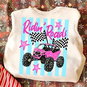 Peut inclure: Sweat-shirt cr&egrave;me avec un motif de v&eacute;hicule tout-terrain rose. Le design comprend le texte "Ridin' Roads" en rose, des rayures verticales bleues, des drapeaux &agrave; damier et des &eacute;toiles roses. Le v&eacute;hicule a de gros pneus noirs.