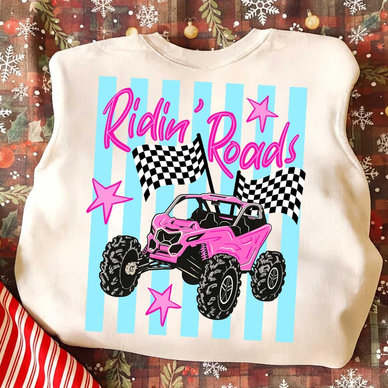 Peut inclure: Sweat-shirt cr&egrave;me avec un motif de v&eacute;hicule tout-terrain rose. Le design comprend le texte "Ridin' Roads" en rose, des rayures verticales bleues, des drapeaux &agrave; damier et des &eacute;toiles roses. Le v&eacute;hicule a de gros pneus noirs.