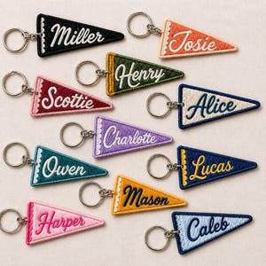 May include: A collection of eleven triangular felt pennant keychains in various colors including black, orange, green, red, cream, purple, teal, dark blue, yellow, pink, and light blue, each featuring embroidered names and a metal keyring.
