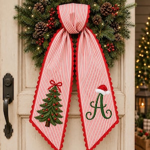 Peut inclure: Une suspension de porte en tissu décoratif avec des rayures verticales rouges et blanches et des bords festonnés rouges. Un côté présente un sapin de Noël vert brodé avec des ornements rouges, l'autre une lettre A verte avec un bonnet de Père Noël.