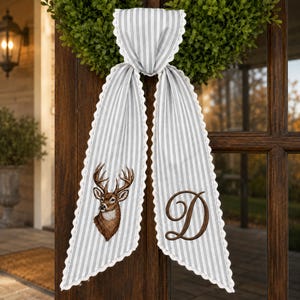 May include: A gray and white striped fabric door hanger featuring a white lace trim along the edges. One side displays a brown deer head illustration, while the other side features a large, brown embroidered letter D in a cursive script font.