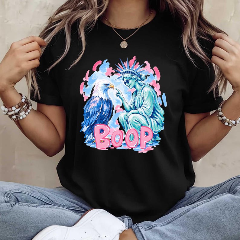 May include: Black t-shirt with a graphic design. The design features a blue and white eagle, a turquoise Statue of Liberty, and the pink word "BOOP." The design is set against a backdrop of pink and blue clouds.
