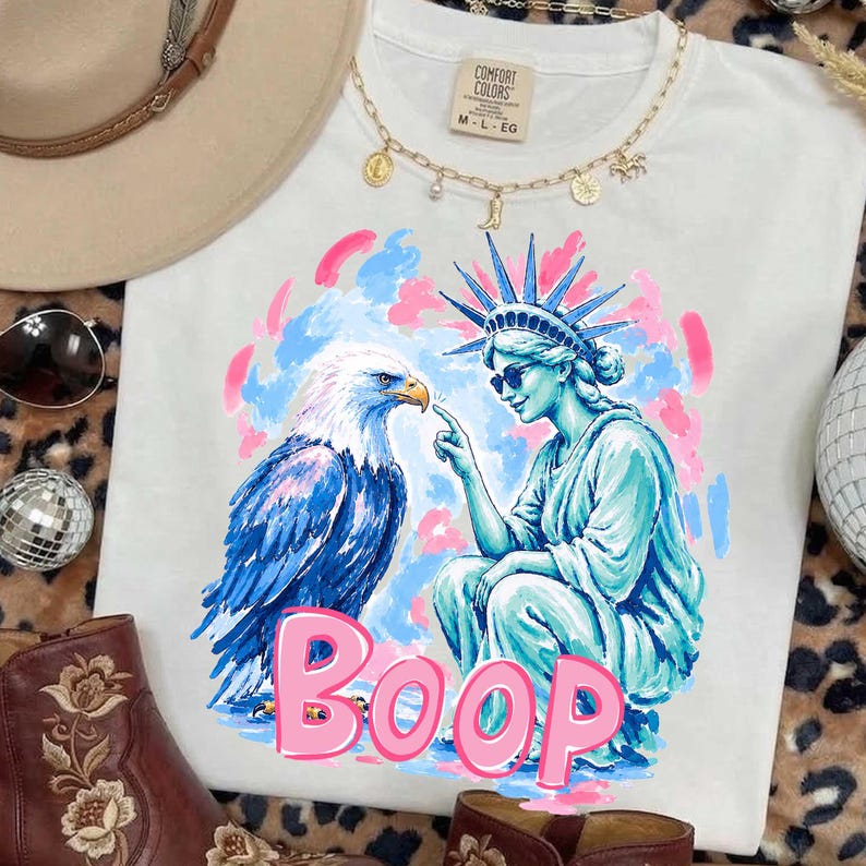 May include: White t-shirt featuring a graphic of the Statue of Liberty and an eagle, with the word "Boop" in pink. The Statue of Liberty is pointing at the eagle. The shirt has a crew neck and short sleeves.