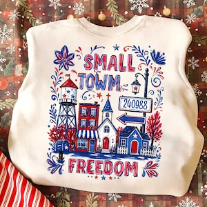 Könnte beinhalten: Cremefarbenes Sweatshirt mit patriotischem Design. Die Grafik zeigt eine Kleinstadt-Szene mit Wasserturm, Gebäuden und den Worten "SMALL TOWN FREEDOM" in Rot und Blau. Ein Straßenschild zeigt die Nummer 240988.