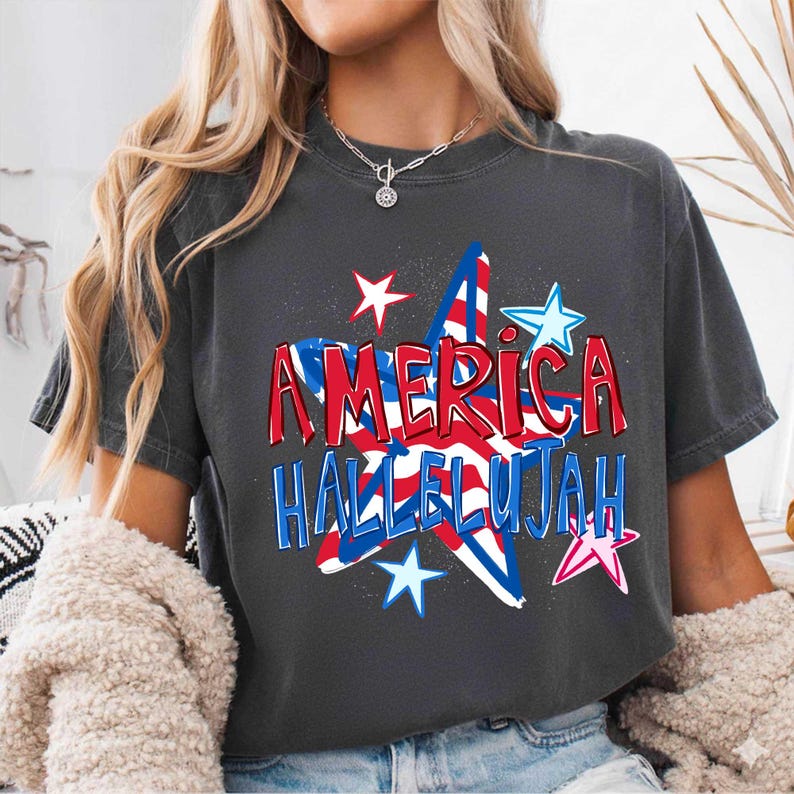 May include: Dark gray t-shirt with the words "AMERICA HALLELUJAH" in red and blue, with a star graphic. The shirt has a vintage, worn look, perfect for patriotic events.