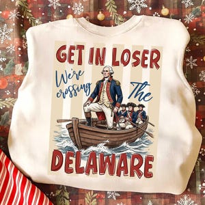 May include: Cream-colored sweatshirt with a graphic of George Washington in a boat crossing the Delaware River. Text reads "GET IN LOSER We're crossing The DELAWARE" in red and blue. The design features a vintage illustration style.