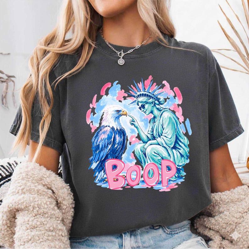 May include: Dark grey t-shirt with a graphic of an eagle, the Statue of Liberty, and the word "BOOP" in pink. The eagle and Statue of Liberty are in blue and white, set against a backdrop of pink and blue paint strokes.