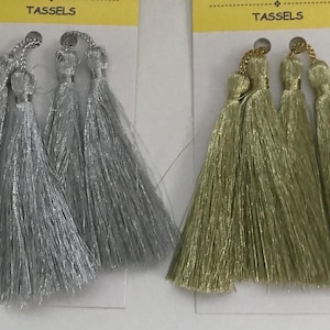 May include: Two sets of decorative tassels attached to white display cards with yellow headers labelled TASSELS. One set features four shimmering silver thread tassels, while the other set features four shimmering gold thread tassels with loops.