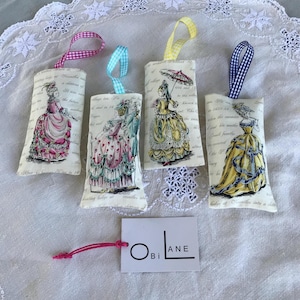May include: Four fabric sachets with a vintage design featuring women in dresses and text. Each sachet has a different colour ribbon and is tied with a bow. The sachets are labelled "Obi Lane".