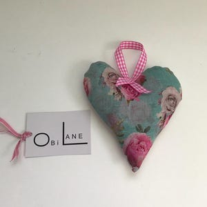 May include: A heart-shaped fabric ornament with a floral pattern in shades of pink and green. The ornament has a pink and white gingham ribbon attached for hanging. The ornament has a tag attached with the text "Obi Lane".