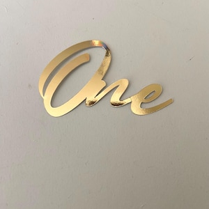 May include: A gold foil sticker with the word "One" in a cursive font. The sticker is a shiny gold colour and is placed on a light grey surface. The sticker is likely used for a birthday or celebration.