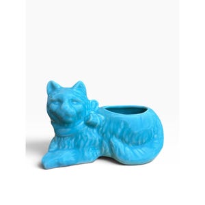 May include: A bright turquoise ceramic planter shaped like a reclining cat. The cat figure features a smooth, glossy finish, molded fur texture, and a decorative bow around its neck. The open basin for plants is located on the cat's back.
