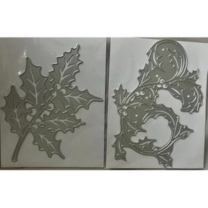 May include: Two silver metal die cuts. One is a maple leaf shape, and the other is a decorative holly design. Both are on a white background. These are likely used for crafting and scrapbooking projects.