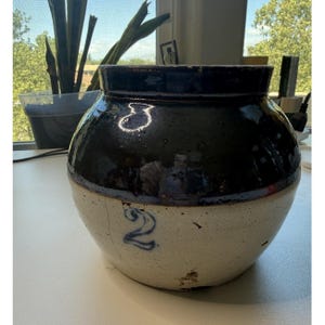 May include: A vintage stoneware crock with a dark blue glazed top and a cream-colored base. The number "2" is painted in blue on the front. The crock has a rounded shape and is likely used for food storage.