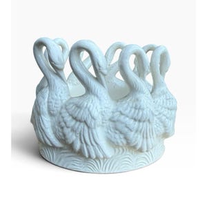 May include: A white ceramic bowl featuring a circular arrangement of sculpted swans around the exterior. The base of the bowl is decorated with a textured, wavy pattern resembling water, while the swan necks curve inward to form the rim.