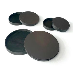 May include: Four black round containers with lids. The containers are made of a smooth, hard plastic. The lids are slightly raised in the center.