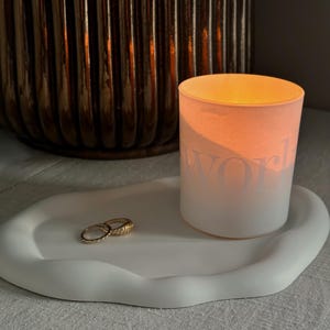May include: A lit candle in a white glass with the word "world" printed on it, on a white, organic-shaped tray. Two gold rings are on the tray. A brown, ribbed vase is in the background.