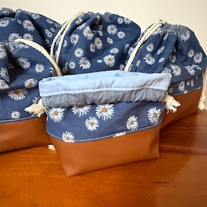 May include: A collection of drawstring bags featuring a denim daisy print and brown faux leather bases. Each bag has a white rope drawstring and white tassels. The bags are displayed on a wooden surface.