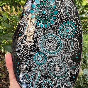 May include: An oval-shaped black stone featuring intricate hand-painted floral and mandala patterns in white and teal. The design includes multiple layered petals, swirling lines, and small dots, accented with embedded iridescent rhinestones.
