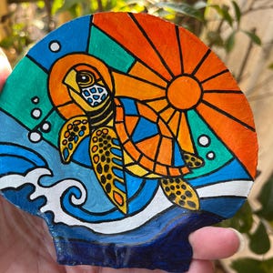 May include: Hand-painted seashell featuring a sea turtle design. The turtle is depicted in vibrant orange, yellow, and blue, with a sunburst background. The artwork includes ocean waves and bubbles, creating a colorful, decorative piece.