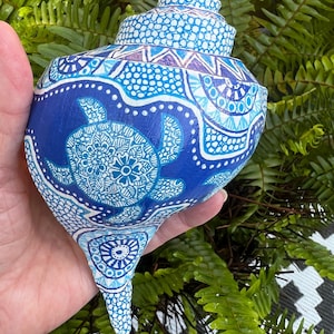 May include: A large spiral seashell painted in shades of blue and white. The surface features intricate hand-painted patterns including a central sea turtle motif, geometric shapes, and fine dot work covering the entire exterior of the shell.