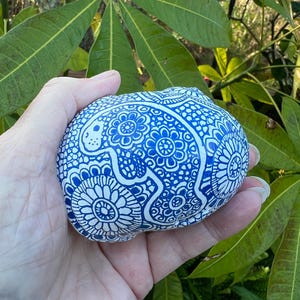 May include: A blue and white painted seashell with a detailed floral and geometric pattern. The shell is held in a hand, with green foliage in the background. The design features intricate, symmetrical elements.