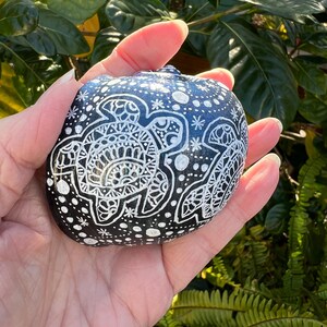 May include: A black and white painted rock with a mandala design of two sea turtles. The rock is covered in white dots and stars.