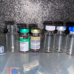 May include: A collection of seven clear glass vials with various colored caps. The vials are arranged in a row on a white surface. The caps are red, green, gold, white, silver, and blue.