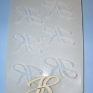 May include: A white silicone mold with six impressions of the letter "R" in a cursive font. A wooden letter "R" is in the foreground.
